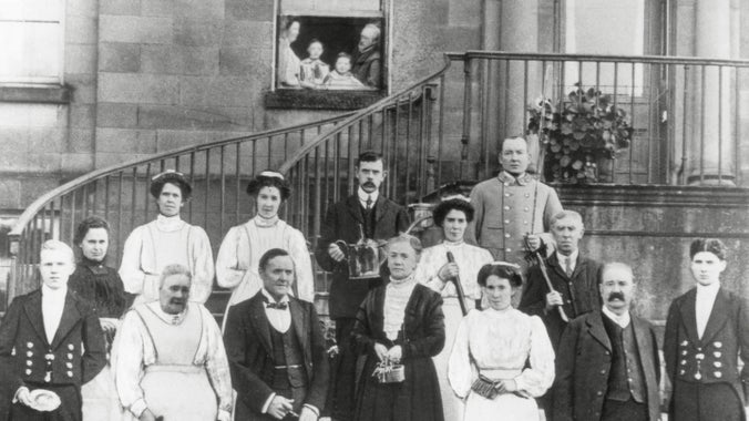 Black and white photo of servants in uniform, standing on the front steps of a grand house. A family of parents and children look on from a first-floor window.